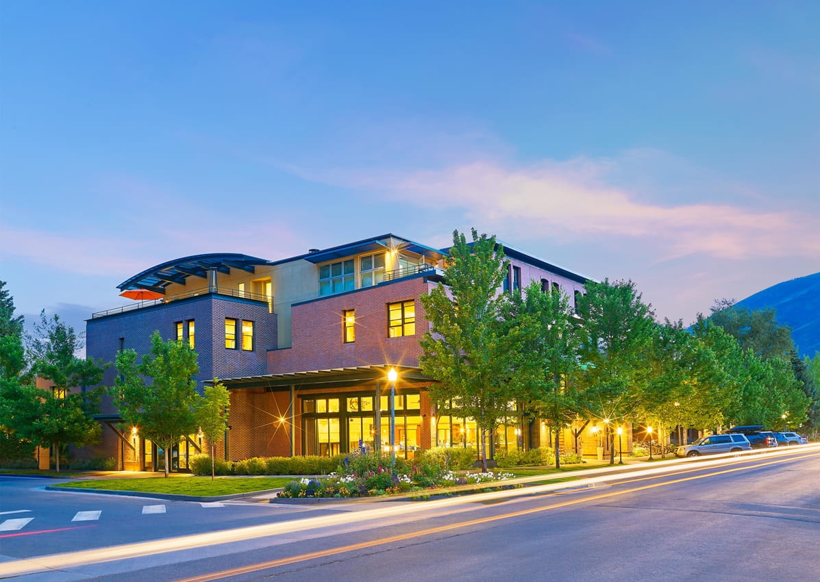 Limelight Aspen resembels a modern brick building with lit windows stands on a tree-lined street at dusk. Parked cars and a distant mountain are visible.